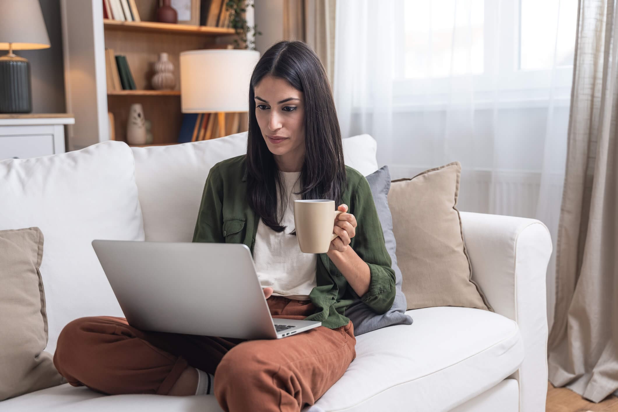 Woman working on laptop while holding a cup of coffee at home.